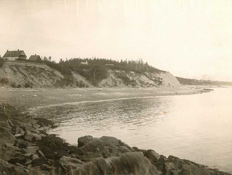 Photographie noir et blanc d’un rivage avec de hautes falaises et, au sommet, une maison