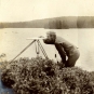Black and white photograph of man standing in front of a lake with surveying equipment