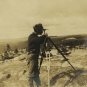 Black and white photograph of man standing on rocks with surveying equipment